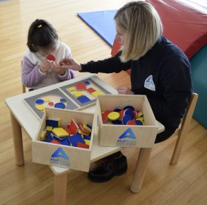 The picture shows an AbilNova rehabilitator working with a blind child on the development of tactile skills, necessary for harmonious development. The skills the child learns at AbilNova are then transferred to the kindergarten with the help of the specialised AbilNova educator, who is present at the school. The child and the rehabilitator are in the AbilNova gymnasium, there are materials for psychomotricity and a small table, suitable for kindergarten children. The girl and the rehabilitator are using wooden geometric shape games, made by AbilNova, to stimulate tactile recognition of shapes, size and thickness. The wooden toys have saturated colours, which also facilitate visual recognition by visually impaired children. 